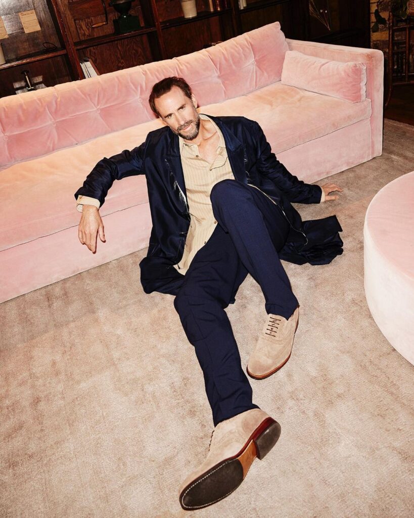 Model wearing a dark suit sitting on the floor with his back against a sofa in the Menier Lounge