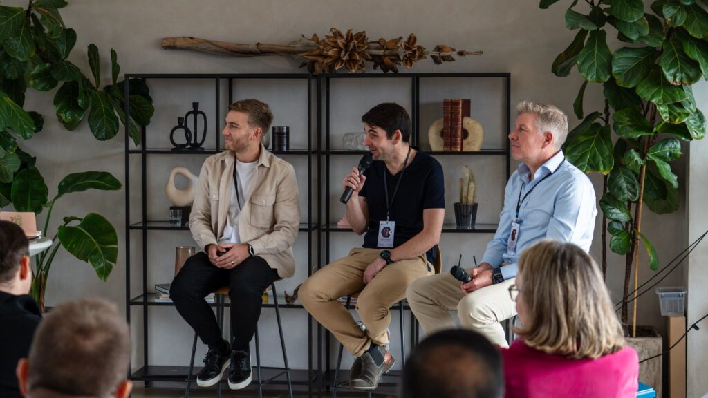 Panel discussion with three male speakers seated on high stools in front of a metal bookshelf during a corporate event.