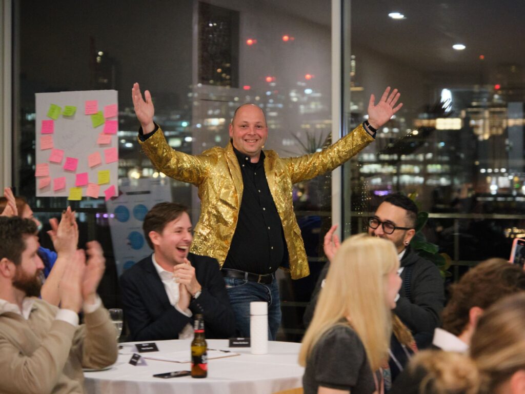 Man in a gold jacket celebrating with arms raised amidst laughing guests at a table during an evening event overlooking London at night.
