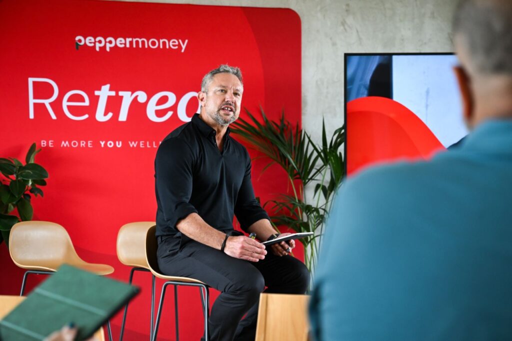 Male host speaking from a stool in front of a red Pepper Money Retreat backdrop during a corporate wellness seminar.