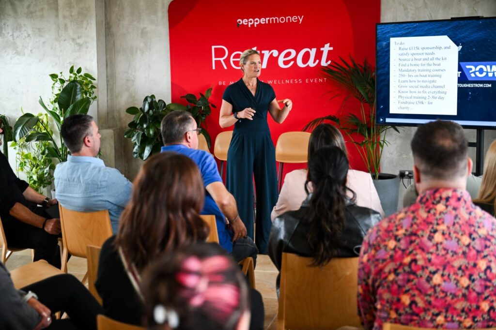 Female speaker presenting to an audience in front of a red backdrop and digital display screen during a wellness workshop.