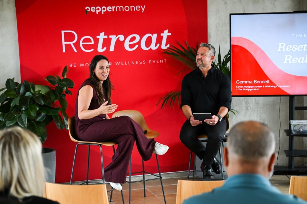 Speaker participating in an fireside chat interview next to a host in front of a red event banner.