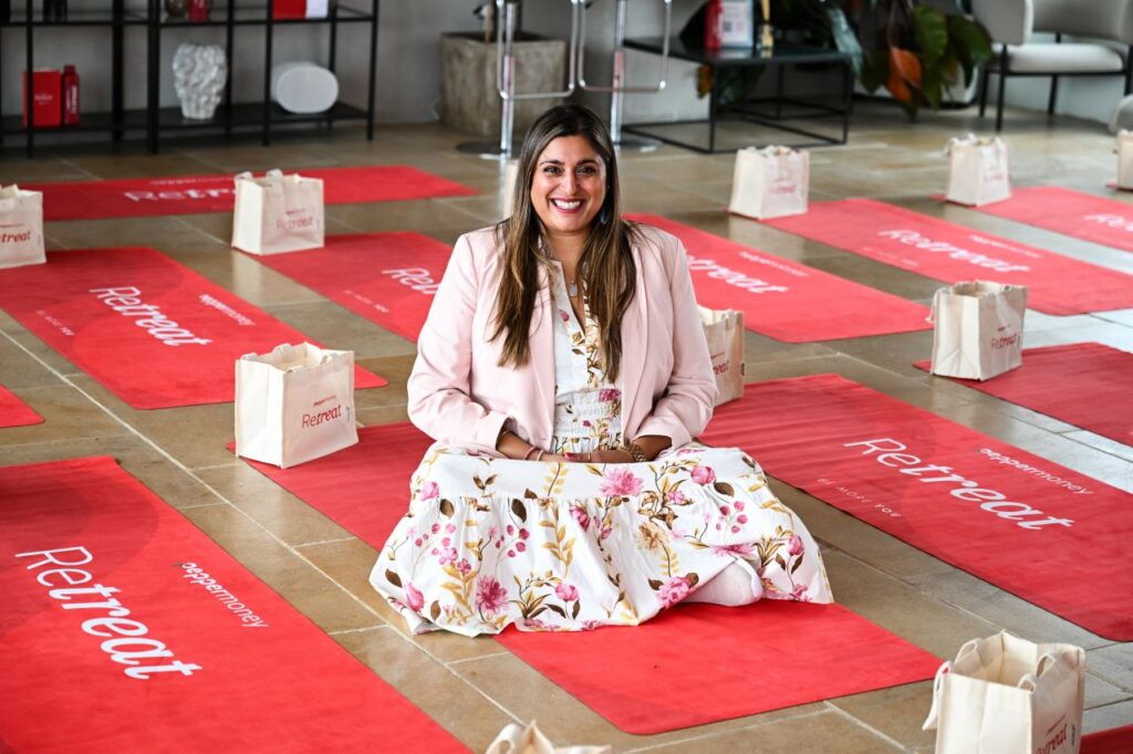 Woman in a floral dress sitting cross-legged on a red yoga mat surrounded by branded mats and gift bags.