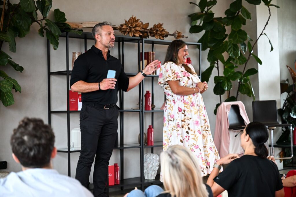Male and female co-hosts standing together addressing an audience seated on yoga mats during a wellness event.