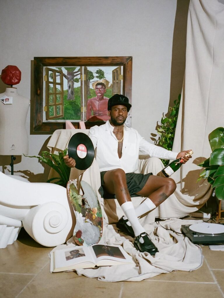 Model seated in a white armchair holding a vinyl record and champagne bottle during an artistic studio photo shoot.