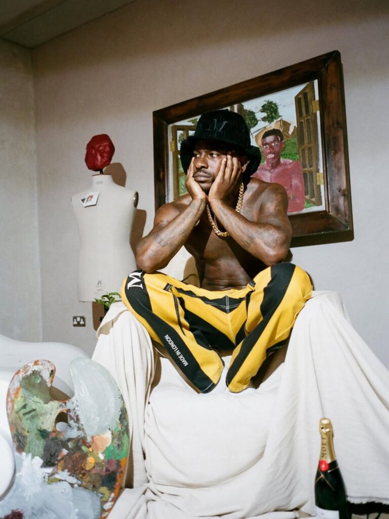 Shirtless man in a black bucket hat and yellow-and-black patterned trousers posing thoughtfully in an artistic studio setting.