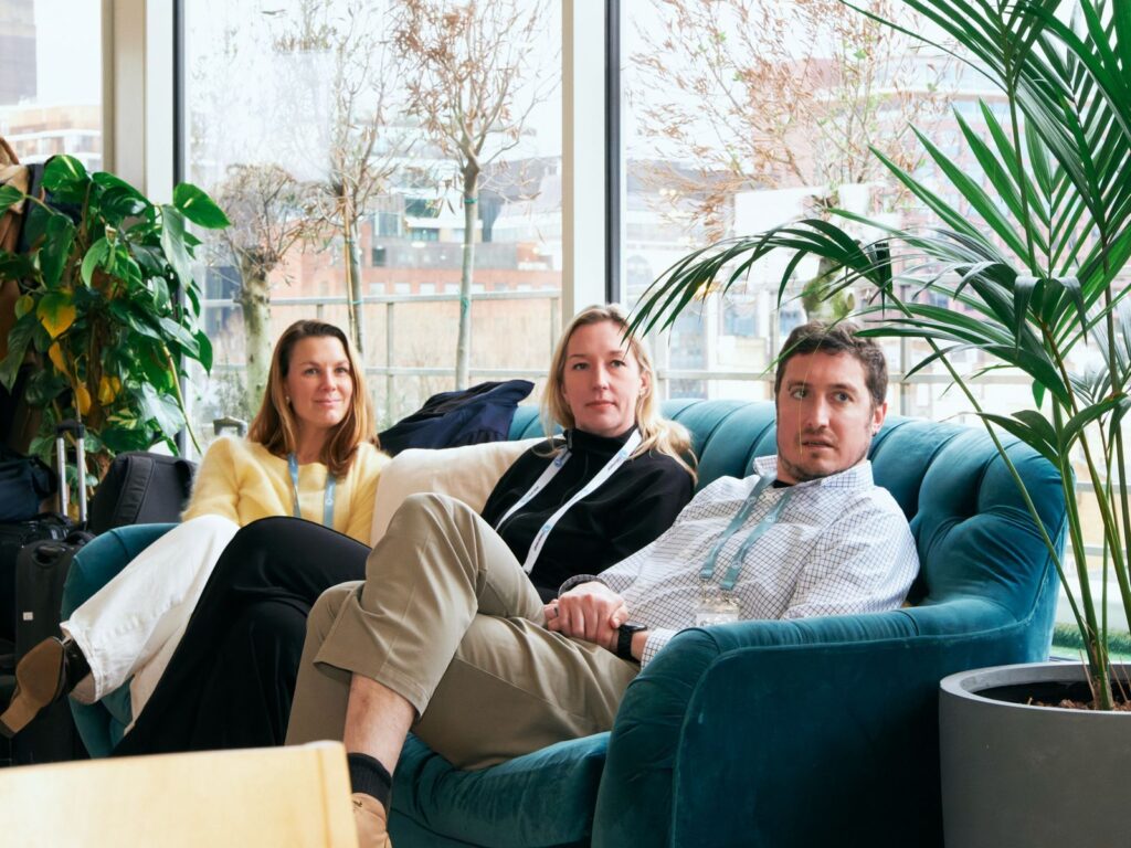 Conference participants sitting on a blue sofa listening to a presentation next to large floor-to-ceiling windows.