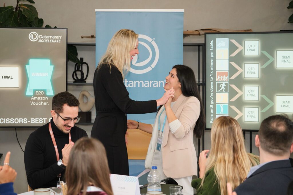 Speaker congratulating an attendee during an interactive workshop exercise on stage in front of Datamaran banners.