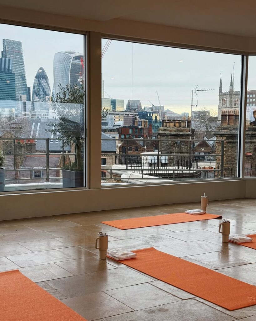 Orange yoga mats laid out on a tiled floor facing floor-to-ceiling windows with views of the London skyline.