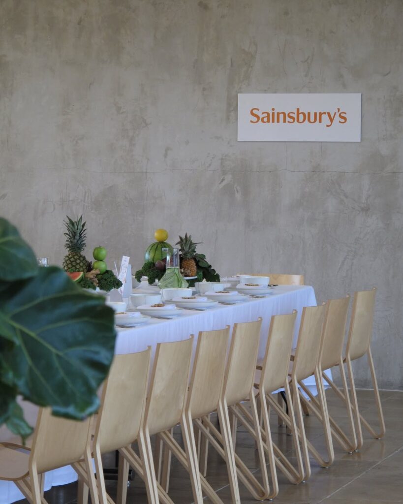Long dining table set with white linen, wooden chairs, and fresh fruit displays below a Sainsbury's sign on a plaster wall.