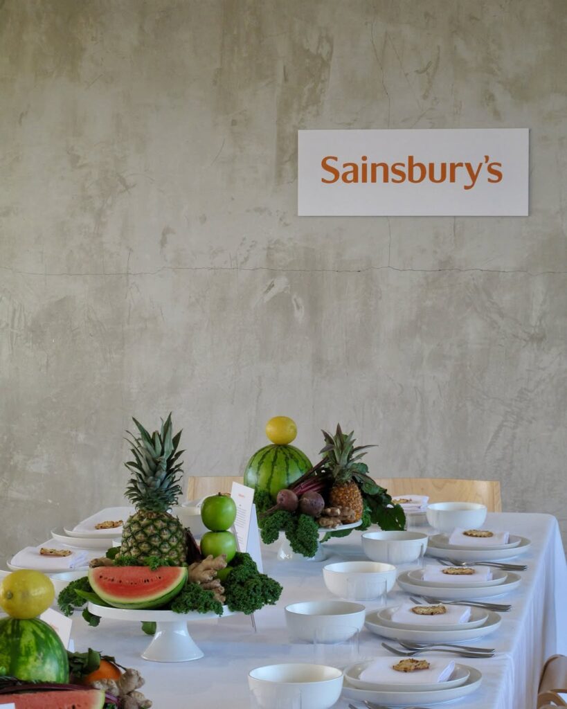 Table arrangement featuring fresh tropical fruit centerpiece displays beneath a Sainsbury's wall sign.